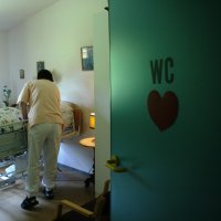 Bildet viser en pasient i en seng på sykehjem, med to pleiere som står ved sengen.