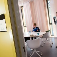 Bildet viser døråpningen til et møterom, der det er to personer. Den ene sitter ved bordet, og den andre står ved siden av og drikker en kopp kaffe.