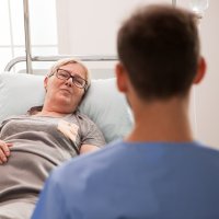 Sykepleier snakker med sengeliggende kvinne.