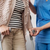 Bildet viser nedre del av kroppene til en dame med krykker og en sykepleier som støtter henne