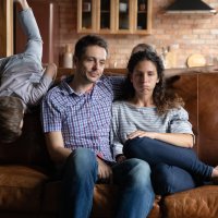 Bildet viser en mann og en kvinne som ser slitne ut og sitter i en sofa, og et barn som klatrer på sofaryggen