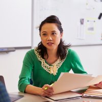 Bildet viser en kvinne som sitter bak et kateter og underviser to eldre elever som vi ser ryggene av