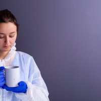 Bildet viser et ungt, kvinnelig helsepersonell i verneutstyr som ser sliten ut og har en kopp i hånden