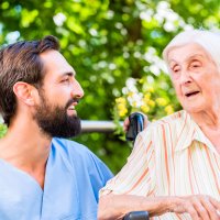 Bildet viser en mannlig sykepleier som snakker med en eldre dame. De sitter ute, og det er god stemning.