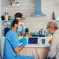 Bildet viser en mannlig sykepleier i blå uniform, munnbind og visir på hjemmebesøk hos en eldre mann, også han med munnbind. De sitter og ser på en ipad.