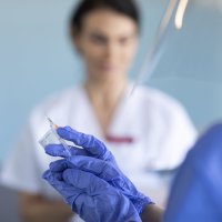 Bildet viser en hånd som holder en sprøyte, og en kvinne i bakgrunnen.