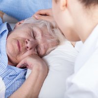 Bildet viser en syk dame i sengen. En yngre dame sitter ved sengekanten og stryker henne over håret