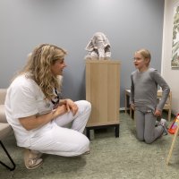 Bildet viser en sykepleier som sitter på huk foran ei jente. De er på et rom på sykehuset med en tøyelefant og et tegnestaffeli.