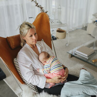 Bildet viser en nybakt mor som sitter i en lenestol hjemme. Hun ser litt oppgitt ut og har babyen liggende i fanget