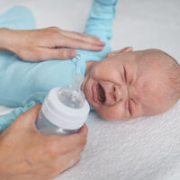 Bildet viser et spedbarn som storgråter. En hånd ligger trøstende på magen til barnet, mens den andre hånden holder en tåteflaske og skal til å gi barnet