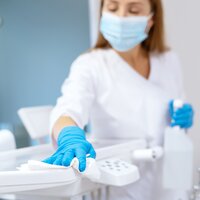 Bildet viser en sykepleier med munnbind og hansker. Hun holder en sprayflaske i hånden og tørker med en fille